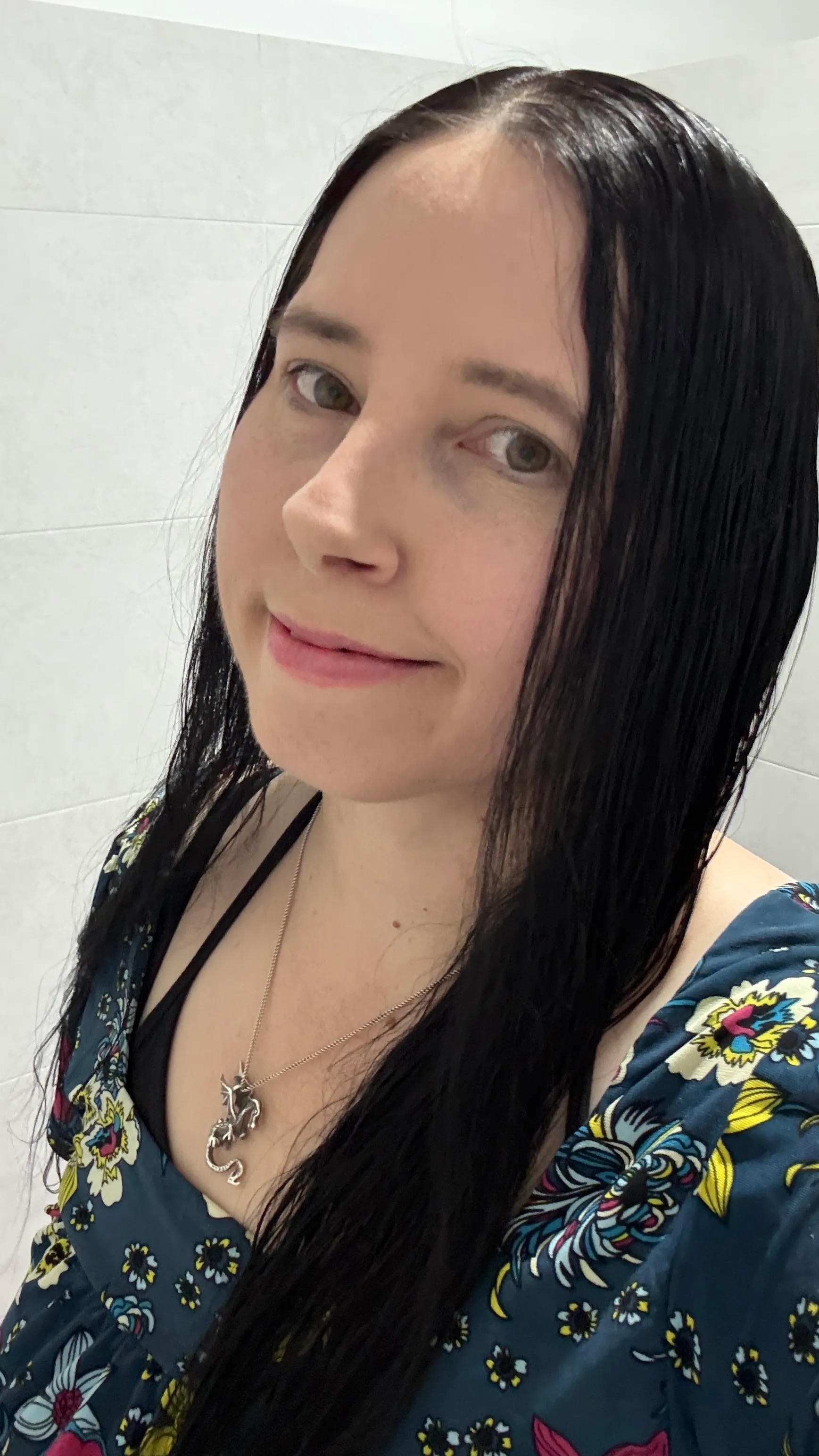 Selfie of Althena Rosalind smiling, with long dark hair, a silver dragon necklace and a teal floral top.